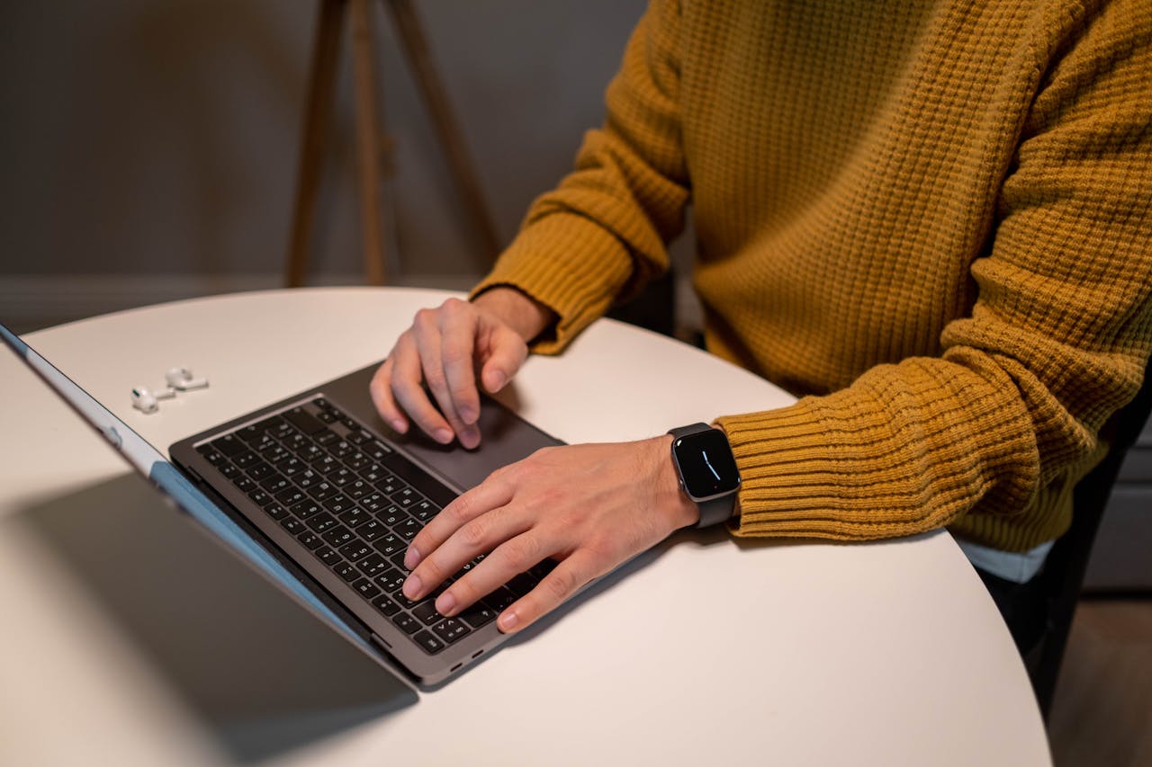Services A man in a yellow sweater uses a laptop and smartwatch at a table. AirPods are visible beside him.